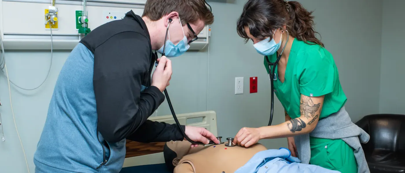 Two physician assistant students use stethoscopes to listen to the heartbeat of a patient simulator